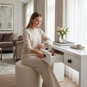 May include: A woman opens a drawer in a white desk. The desk has black knobs and a tray with a cup and saucer. A vase of flowers and a book are on the desk. The woman is wearing a cream-colored sweater and pants.