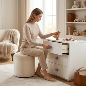 May include: A white dresser with purple knobs, a wooden duck figurine, and a beige ottoman. The dresser has multiple drawers and is in a well-lit room. A person is opening a drawer. The room also features a bookshelf and a chair.