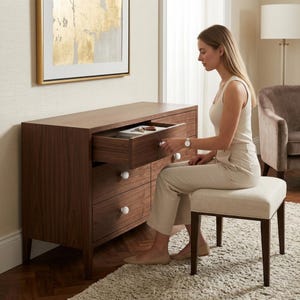 May include: A dark brown wooden dresser with six drawers, featuring white round knobs. The top drawer is open, revealing items inside. A woman is seated on a beige upholstered stool in front of the dresser. The room has a neutral color palette.
