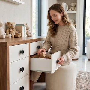 May include: A woman opening a white drawer with a black knob on a wooden dresser. The dresser has a brown top and white drawers. Teddy bears and books are on the top of the dresser. The room has a neutral color palette.