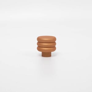 Puede incluir: Un pomo de armario de color bronce con un dise&ntilde;o apilado y redondeado. El pomo presenta tres segmentos circulares horizontales sobre una base cil&iacute;ndrica m&aacute;s peque&ntilde;a. El acabado tiene un brillo met&aacute;lico y el pomo est&aacute; sobre un fondo blanco liso.