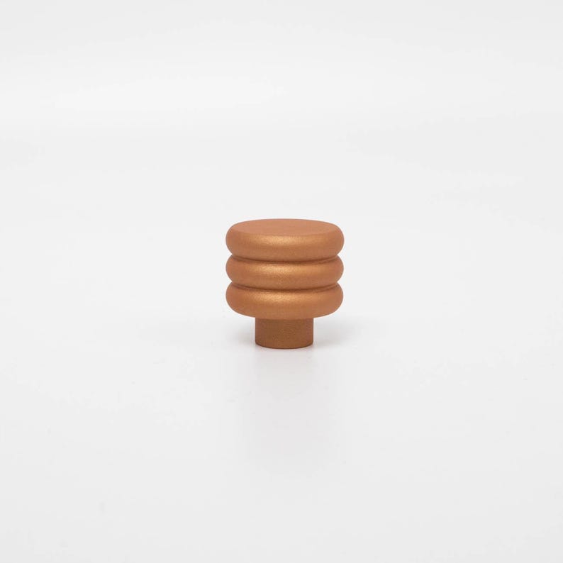 Puede incluir: Un pomo de armario de color bronce con un dise&ntilde;o apilado y redondeado. El pomo presenta tres segmentos circulares horizontales sobre una base cil&iacute;ndrica m&aacute;s peque&ntilde;a. El acabado tiene un brillo met&aacute;lico y el pomo est&aacute; sobre un fondo blanco liso.