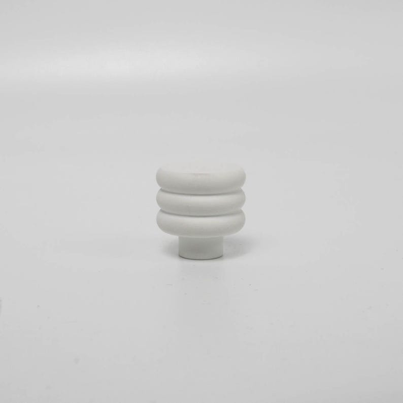 Puede incluir: Un pomo de armario blanco con un dise&ntilde;o apilado y redondeado. El pomo presenta tres segmentos horizontales en forma de anillo sobre una base cil&iacute;ndrica corta. El acabado liso y mate le da una est&eacute;tica moderna. Ideal para mejoras de muebles.
