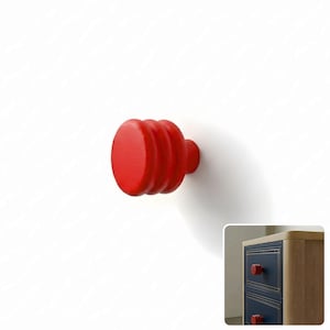 May include: A bright red wooden drawer knob with a cylindrical shape and three distinct horizontal ridges along its side. The knob features a flat circular front face and is shown installed on a dark blue wooden drawer unit with light wood trim.