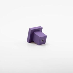 K&ouml;nnte beinhalten: Ein quadratischer Schrank-Knopf in Lila mit einem Schraubenloch an der Unterseite.
