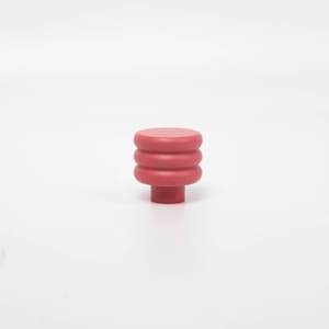 Puede incluir: Un pomo de armario rojo liso con un dise&ntilde;o apilado y redondeado. El pomo presenta una base cil&iacute;ndrica y tres secciones apiladas en forma de anillo que conducen a una parte superior plana y circular. El color es un rojo apagado.