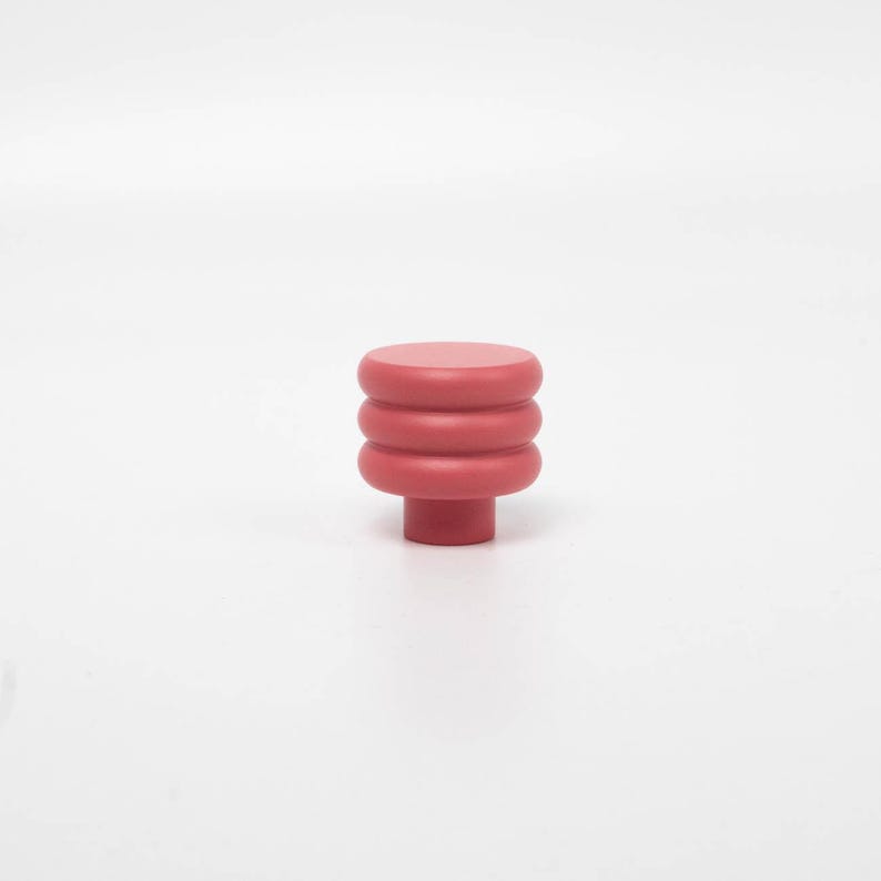 Puede incluir: Un pomo de armario rojo liso con un dise&ntilde;o apilado y redondeado. El pomo presenta una base cil&iacute;ndrica y tres secciones apiladas en forma de anillo que conducen a una parte superior plana y circular. El color es un rojo apagado.