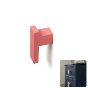May include: A coral pink T-shaped cabinet knob with a smooth matte finish. The knob features a rectangular base and a protruding handle. A smaller inset image shows the same coral pink knob installed on a dark navy blue wooden drawer unit.