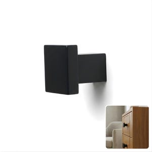 May include: A matte black wall hook featuring a square-shaped front plate and a rectangular base. The hook has a clean, geometric design with sharp edges and a solid, uniform finish. A small inset shows the hook installed on a wooden cabinet drawer.