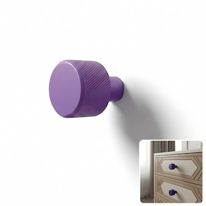 May include: A purple cabinet knob with a cylindrical shape and a textured, diagonal ribbed pattern on its side. The knob features a smooth, flat circular front face and a smaller cylindrical base that attaches to a drawer or cabinet door surface.