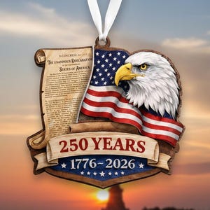 May include: A wooden ornament featuring an American flag, a bald eagle, and a scroll with text. The ornament reads "250 YEARS 1776-2026" and is suspended by a white ribbon. The background shows a sunset over water.