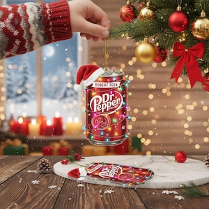 May include: A red Dr. Pepper can-shaped Christmas ornament with a Santa hat and string lights. The ornament has the text "ROBERT 2024" and "Dr. Pepper Fryer" on it. A second ornament lies on a marble surface.