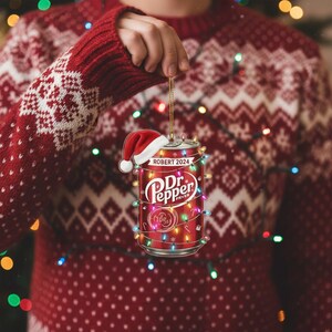May include: A red Dr. Pepper can Christmas ornament with a Santa hat and string lights. The ornament has the text "ROBERT 2024" and the Dr. Pepper logo. The ornament is being held by a person wearing a red Christmas sweater.