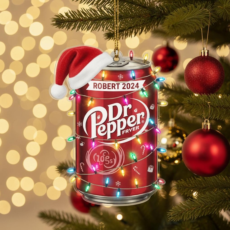 May include: A red Dr. Pepper can Christmas ornament with a Santa hat, string lights, and the text "ROBERT 2024". The ornament is hanging on a Christmas tree with red ornaments.