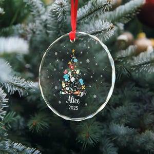 May include: A clear, round Christmas ornament with a red ribbon. The ornament features a Christmas tree design made of medical supplies and the name "Alice" and the year "2025".