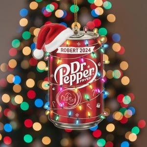 May include: A red Dr. Pepper can Christmas ornament with a Santa hat and string lights. The ornament has the text "ROBERT 2024" and the Dr. Pepper logo. The can is decorated with colorful Christmas lights.