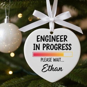 May include: White heart-shaped Christmas ornament with the text "ENGINEER IN PROGRESS" and "PLEASE WAIT..." in black. A progress bar is displayed below the text. The name "Ethan" is written at the bottom. The ornament is tied with a white ribbon.