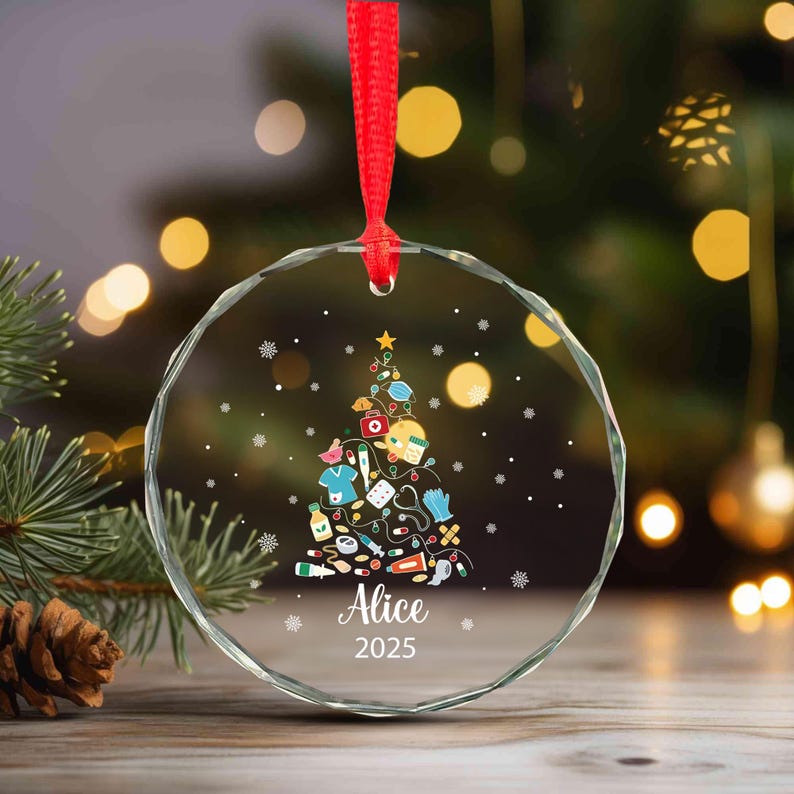 May include: A clear, round Christmas ornament with a red ribbon. The ornament features a Christmas tree design made of medical supplies and a star on top. The name "Alice" and the year "2025" are printed on the ornament.