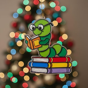 May include: A whimsical Christmas ornament featuring a green caterpillar wearing glasses and reading a book. The caterpillar sits atop a stack of colorful books. The ornament is suspended by a gold string, with a blurred Christmas tree background.