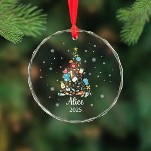 May include: A clear, round Christmas ornament with a red ribbon. The ornament features a Christmas tree design made of medical supplies, a star, and snowflakes. The name "Alice" and the year "2025" are printed on the ornament.