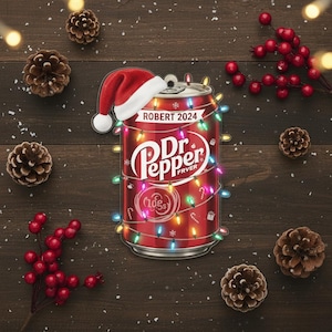 May include: A red Dr. Pepper can-shaped ornament with the text "ROBERT 2024" and the Dr. Pepper logo, wrapped in Christmas lights and topped with a Santa hat. The ornament is surrounded by pine cones and red berries.