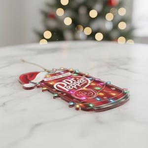 May include: A red Dr. Pepper can-shaped Christmas ornament with a Santa hat and string lights. The ornament features the Dr. Pepper logo and the text "ROBERT TEXAS". The ornament is on a white marble surface.