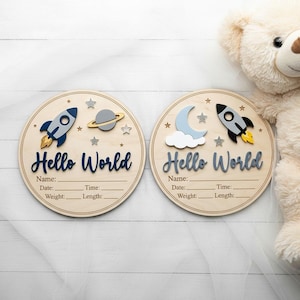 May include: Two circular wooden birth announcement signs featuring the text Hello World. The left sign displays a rocket and planet with stars. The right sign features a moon, cloud, and rocket. Both include lines for Name, Date, Time, Weight, and Length.