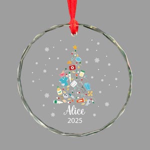 May include: A clear glass Christmas ornament with a red ribbon. The ornament features a Christmas tree design made of medical supplies and a star on top. The name "Alice" and the year "2025" are printed on the ornament.