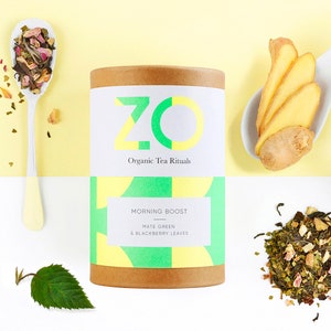 Peut inclure: Un tube en carton brun avec une étiquette blanche qui indique "ZO Organic Tea Rituals" et "Morning Boost Mate Green & Blackberry Leaves". L'étiquette présente un motif géométrique vert et jaune. Le tube est posé sur une surface blanche.