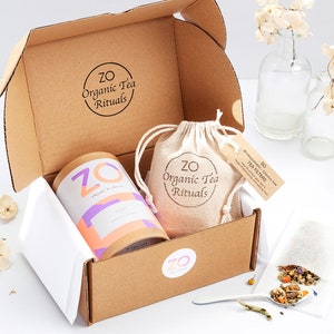 May include: A brown cardboard box with a white tissue paper lining. The box contains a round container of organic tea labelled "ZO Sleep", a small burlap bag with the text "ZO Organic Tea Rituals", and a small bag of tea filters. The box is open and the contents are visible.