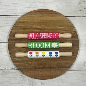 May include: Three wooden rolling pins with spring-themed designs. The top rolling pin is red with the text "HELLO SPRING" and a butterfly. The middle rolling pin is green with the text "BLOOM" and a daisy. The bottom rolling pin is white with a row of colorful tulips.