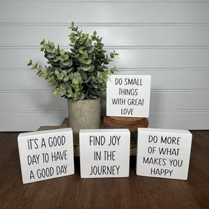 May include: Four white wooden blocks with inspirational quotes. The quotes are: "It's a good day to have a good day", "Find joy in the journey", "Do more of what makes you happy", and "Do small things with great love".
