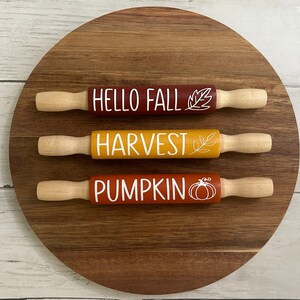 May include: Three wooden rolling pins with fall-themed designs. The top pin is red and says "HELLO FALL" with a leaf design. The middle pin is yellow and says "HARVEST" with a leaf design. The bottom pin is orange and says "PUMPKIN" with a pumpkin design.