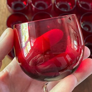 May include: A set of small, round, red glass cups. The cups are clear and have a slight reddish tint. They are arranged in a row, with one cup being held in a hand.