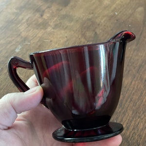 May include: A deep red glass creamer with a footed base and a handle. The creamer is made of glass and has a shiny finish.