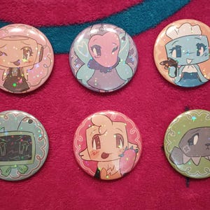 Pode incluir: Seis botões redondos com personagens de desenhos animados coloridos. Os personagens são uma fada rosa, um cozinheiro azul, um robô marrom, uma criatura amarela, um monstro verde e uma criatura marrom com uma estrela na cabeça.