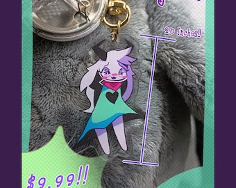 May include: A 2.0 inch clear acrylic keychain featuring a cartoon character with purple hair, a green dress, and a pink heart on a black background. The text "2.0 Inches!" is visible in the image. The keychain is on a gray fuzzy surface.