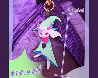 May include: A 2.5 inch keychain featuring a cartoon character with pink hair, a green dress, and a red bandana. The character is holding a wand and has a surprised expression. The keychain is attached to a gold metal chain and is hanging from a purple fuzzy surface. The text "Ralsei 2.5 Inch Keychain" and "$12.99!!" are visible in the image.