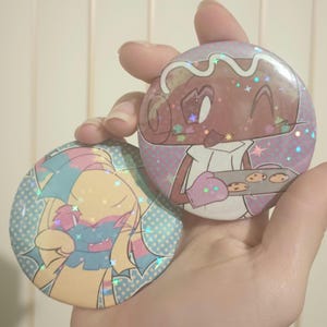 Pode incluir: Dois pinos redondos com acabamento brilhante. Um pino apresenta um personagem de gengibre de desenho animado segurando uma assadeira com biscoitos. O outro pino apresenta um personagem de desenho animado com pelo azul e amarelo. Ambos os pinos têm um fundo de bolinhas azuis e brancas.