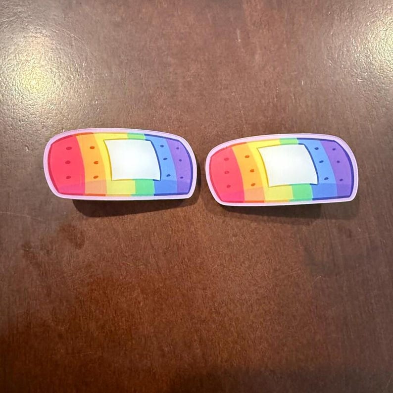 May include: Two decorative band-aids with a rainbow color scheme. Each band-aid has a pink border, a white center, and a rainbow gradient from red to purple. The band-aids are designed to look like a bandage.