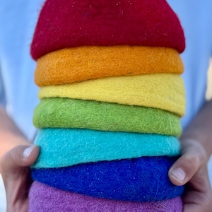 Puede incluir: Una pila de siete cuencos de lana afieltrada en colores del arcoíris: rojo, naranja, amarillo, verde, turquesa, azul y morado. Los cuencos están sujetos por las manos de una persona. Los cuencos tienen unos 10 cm de diámetro.