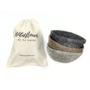 Op de afbeelding: Een set van drie vilten kommen in grijstinten en bruin, op elkaar gestapeld. Naast de kommen staat een natuurlijke canvas tas met de tekst "Wildflower by hu hands". Het websiteadres www.huhands.com staat op de tas.