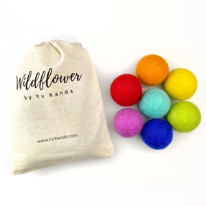May include: A natural canvas bag with the text "Wildflower by hu hands" next to seven colorful felt balls. The balls are in shades of red, orange, yellow, green, blue, teal, and purple. The website address www.huhands.com is printed on the bag.