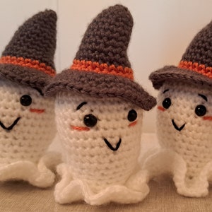 Peut inclure: Trois ornements fantômes en crochet avec des chapeaux de sorcière gris et des bordures orange. Les fantômes ont des corps en crochet blanc et des yeux noirs avec des joues roses.