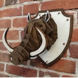 May include: A wooden wall mount sculpture of a warthog head with large white tusks. The sculpture is mounted on a white shield-shaped plaque with silver metal accents.