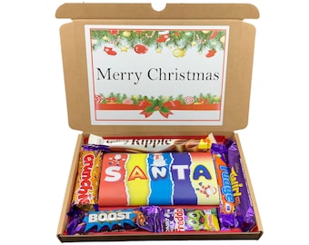 May include: A brown cardboard box with a Merry Christmas message on the lid. The box is open and filled with various chocolate bars, including Galaxy Ripple, Crunch, Boost, Freddo, and Twirl. The chocolate bars are arranged to spell out the word "SANTA".