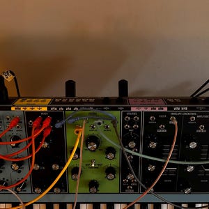 Pode incluir: Um sintetizador modular com vários módulos coloridos, botões e cabos de conexão. Os módulos são em tons de cinza, verde e roxo. O sintetizador está acima de um teclado, com a marca "Moog" visível.