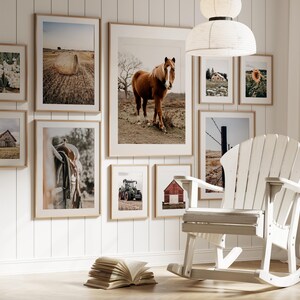 May include: A white rocking chair with a wooden frame sits in front of a wall with framed pictures of horses, barns, and farm scenes. The pictures are of various sizes and are arranged in a grid pattern.