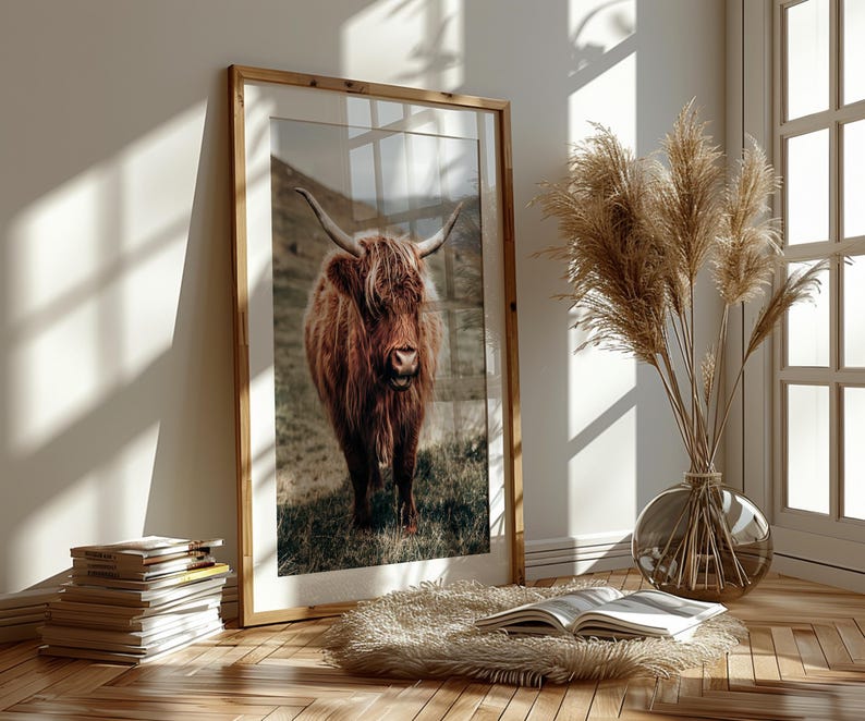 May include: A framed photograph of a brown Highland cow standing in a field of green grass. The cow has long, shaggy hair and a white face. The photograph is in a wooden frame.