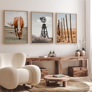 May include: Three framed prints on a white wall above a wooden table. The prints feature a brown horse grazing in a field, a windmill in a field, and a wooden fence in a field. The table has a wooden box on the left side and a wooden box on the right side. There is a small round wooden table in front of the table with a book on top.